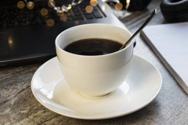 Beyaz bir fincan kahve, tahta bir masanın üzerinde Bokeh ve Notepad ile dizüstü bilgisayarın arka planında duruyor. İş sırasında ara verme ve iş toplantısı kavramı.