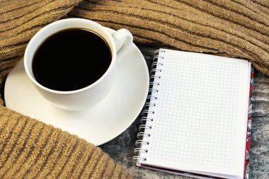 Beyaz bir kahve fincanı ve açık bir not defteri. Yazı için yeri var. Tahta bir masanın üzerinde, süvetere sarılı..