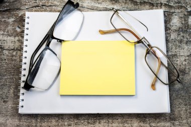 Tahta yüzeyde üzerinde notlar için sarı bir kağıt ve bir çift eski gözlük olan bir not defteri var..