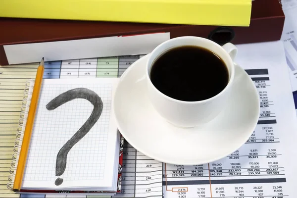On documents with numbers is a Notepad with a question mark on it. A white Cup of coffee next to a folder of documents.