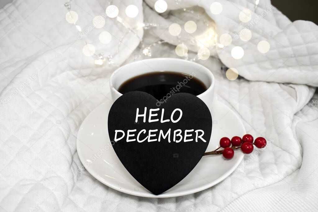 Hello December is written on a black heart that stands on a white saucer near a white Cup of coffee.White background with bokeh.
