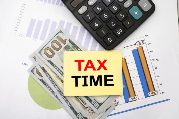 The time of taxes is written on a piece of paper that lies on financial documents with a diagram and paper bills in the form of dollars near the calculator. Business and financial concept