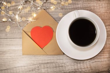 İçinde kırmızı bir kalp olan açık bir zarf. Beyaz bir kahve bardağının yanında. Tahta bir masada. Şenlik sabahı konsepti