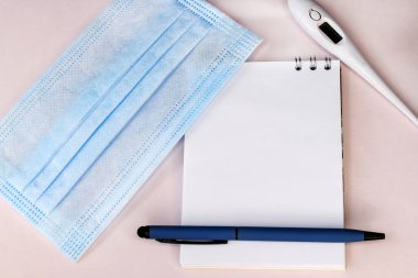 Pembe arka planda, yanında tıbbi yüz maskesi ve elektronik termometre olan boş bir defter var. Tıbbi konsept