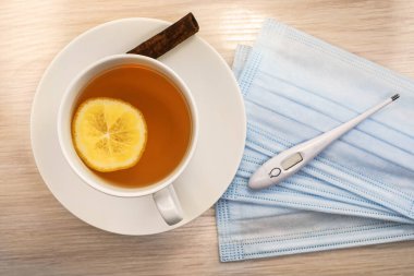 Beyaz bir bardak limon çayı ahşap bir masada bir yığın tıbbi yüz maskesi ve termometrenin yanında duruyor. Tıbbi konsept