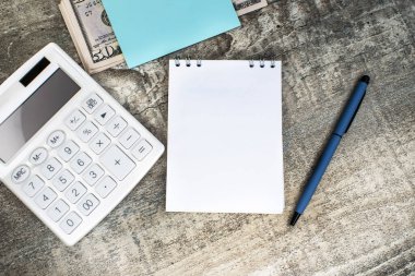 Tahta bir masanın üzerinde beyaz bir hesap makinesi ve dolarların arasında beyaz boş bir sayfası olan bir defter var. İş ve finansal konsept. İş Şablonu