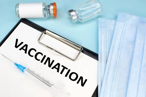 Aşı kelimesi bir kırtasiye tabletinde yazılı. Tıbbi yüz maskesi ve şırıngalı ampuller. Tıbbi konsept