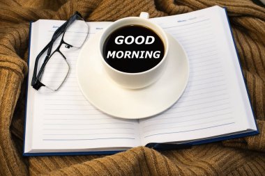 Üzerinde günaydın yazan beyaz bir kahve fincanı. Not defteri ve gözlüklü ceket.