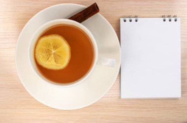 Ahşap masada beyaz bir fincan limon çayı ve boş beyaz çarşaflı bir defter var..