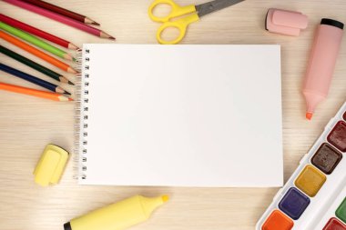 Okul malzemelerinin arasındaki ahşap masanın üzerinde boş beyaz bir çarşaf olan bir defter var. Makas ve kalemle boyanmış renkli kalemler. Şablon ve afiş. Eğitim