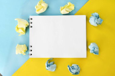 Beyaz boş bir sayfa içeren bir defter, buruşmuş kağıt sayfaları arasında iki renkli bir zemin üzerinde yatar..