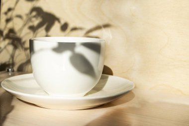 Ahşap yüzeyde beyaz bir kahve fincanı ve üzerine düşen bir bitkinin gölgesi olan bir fincan tabağı var.