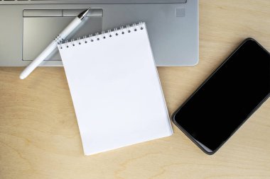 Üzerinde beyaz boş bir sayfa olan bir defter siyah bir telefonun yanındaki gri bir dizüstü bilgisayarın üzerinde duruyor. İş kavramı ve çevrimiçi eğitim