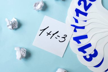 Plastik bir fanın yanında, üzerinde sayılar ve buruşuk kağıtlar olan mavi bir fonda, üzerinde yanlış bir çözüm sonucu olan bir örneğin yazıldığı bir sayfa var. Eğitici kavram