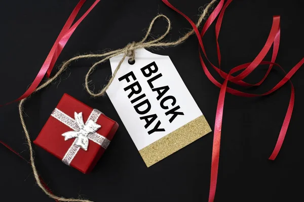 Black Friday is written on the tag for the price tag which lies on a black background near the gift box and a red ribbon. The concept of discounts and sales