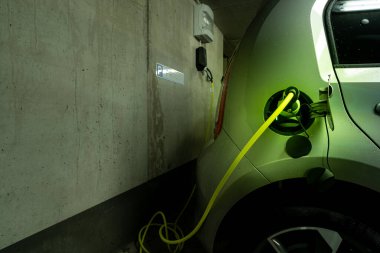 Arka planında ev şarj istasyonu olan elektrikli bir arabanın şarj prizinde, elektrik tip 2 kablosu takılı. Arabanıza yakıt ikmali için trend yaşam tarzı kavramı.