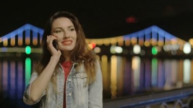 Şehrin arka plan köprü aydınlatıcı ve nehirde su yansımalar ile gece telefonda konuşurken kız
