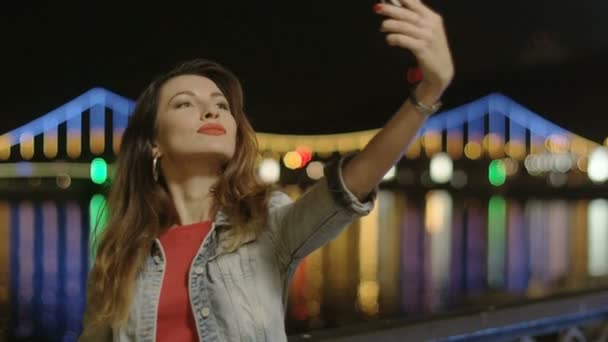 Mulher engraçada bonita que faz selfie com um telefone na cidade da noite no rio com reflexos da água — Vídeo de Stock