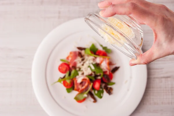 Woman pours a glass plate of cheese in a salad of tomatoes and h — Stock Photo, Image