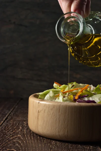 Minyak zaitun mengalir dari kendi kaca ke dalam salad sayuran di kayu — Stok Foto