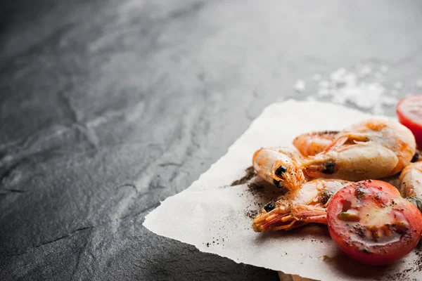 Gamberetti alla griglia sulla pace della carta con pomodori e pepe sul tavolo di pietra nera — Foto Stock