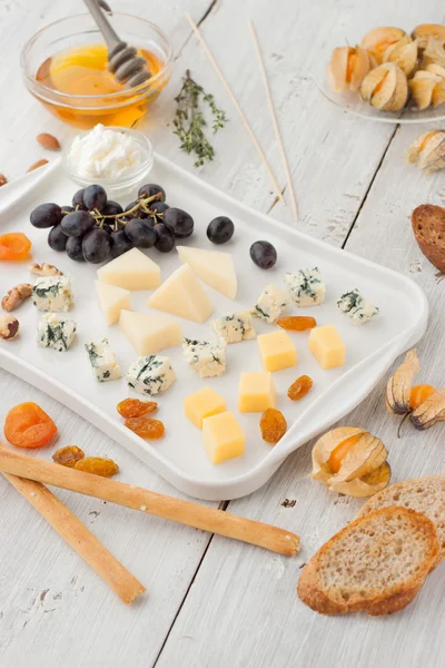 Prato de queijo com frutas e nozes diferentes na mesa de madeira branca vertical — Fotografia de Stock