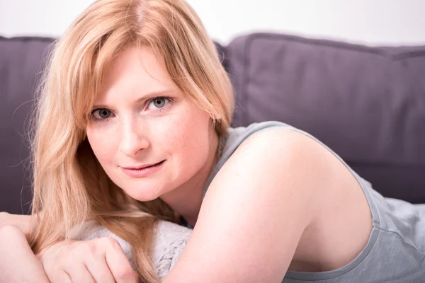 Beautiful blonde woman is smiling and relaxing on gray couch — Stock Photo, Image