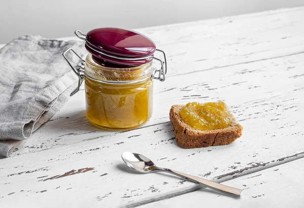 A glass jar of homemade jam, a toast with jam and a spoon on white wooden table, concept of healthy organic breakfast