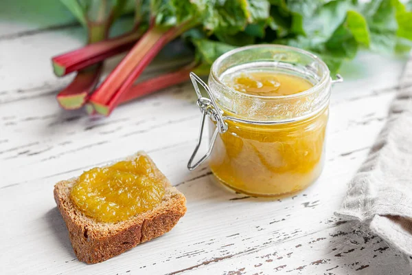 A toast with homemade rhubarb jam for a healthy breakfast, an opened glass jar of jam and fresh rhubarb stalks on a white wooden table