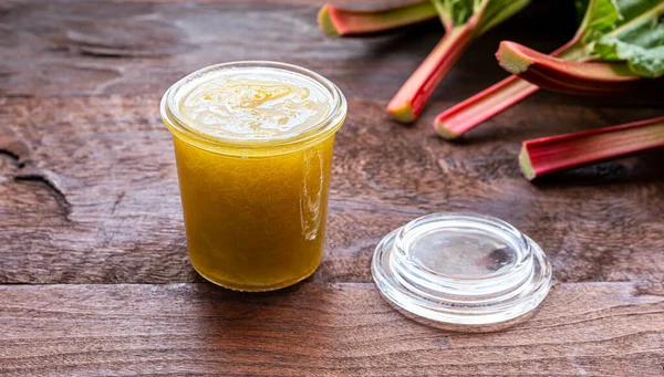 Homemade rhubarb jam for healthy breakfast in a glass jar and fresh rhubarb stalks on a dark old wooden table