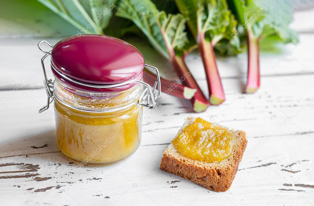 A glass jar of rhubarb jam, a toast with homemade jam for a healthy breakfast and fresh rhubarb stalks on a white wooden table