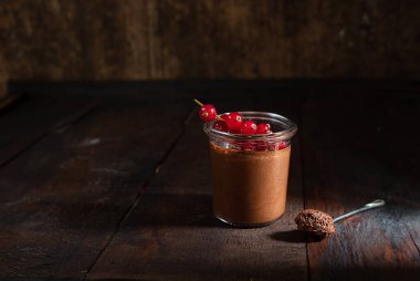 Çikolatalı kremalı bir çay kaşığı, çikolatalı bir bardak ve siyah ahşap masada kırmızı frenk üzümü.