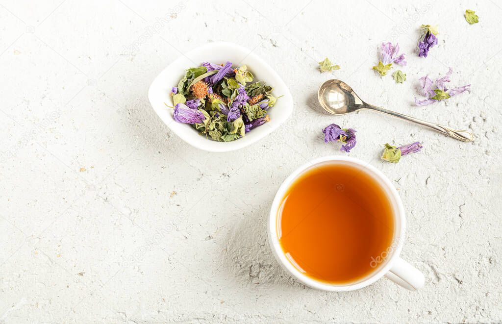 A white cup and a bowl with herbal tea - thyme, green anise, ground ivy, pine bud and mallow flowers on a light gray table, top view with copy space