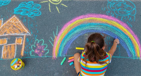 The child draws a house and a rainbow on the asphalt with chalk. Selective focus. Kids.