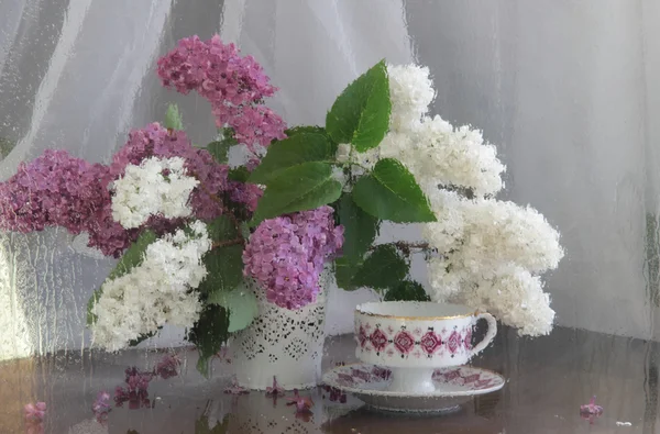 Nature morte avec un lilas et une tasse de thé derrière un verre mouillé — Photo