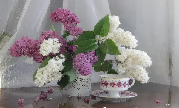 Nature morte avec un lilas et une tasse de thé derrière un verre mouillé — Photo