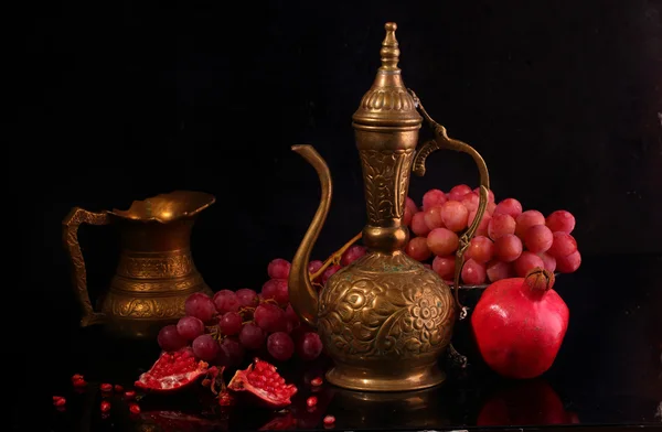 Медный кувшин, красный гранат и розовый виноград на черной спине — стоковое фото