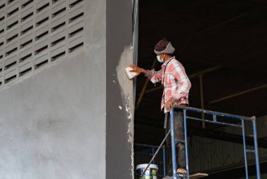 Yüksek duvarlı bir fabrikada boyanmaya hazırlanırken yeni boyanmış gri bir duvarın yüzeyini sıva ile zımparalıyor..