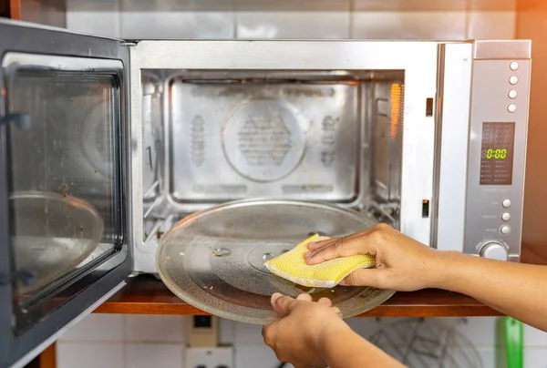 Woman cleaning turn disk in microwave oven with sponge.
