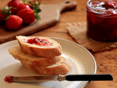 Sliced bread and knife on plate, raw strawberries on cutting board, homemade jan in glass jar