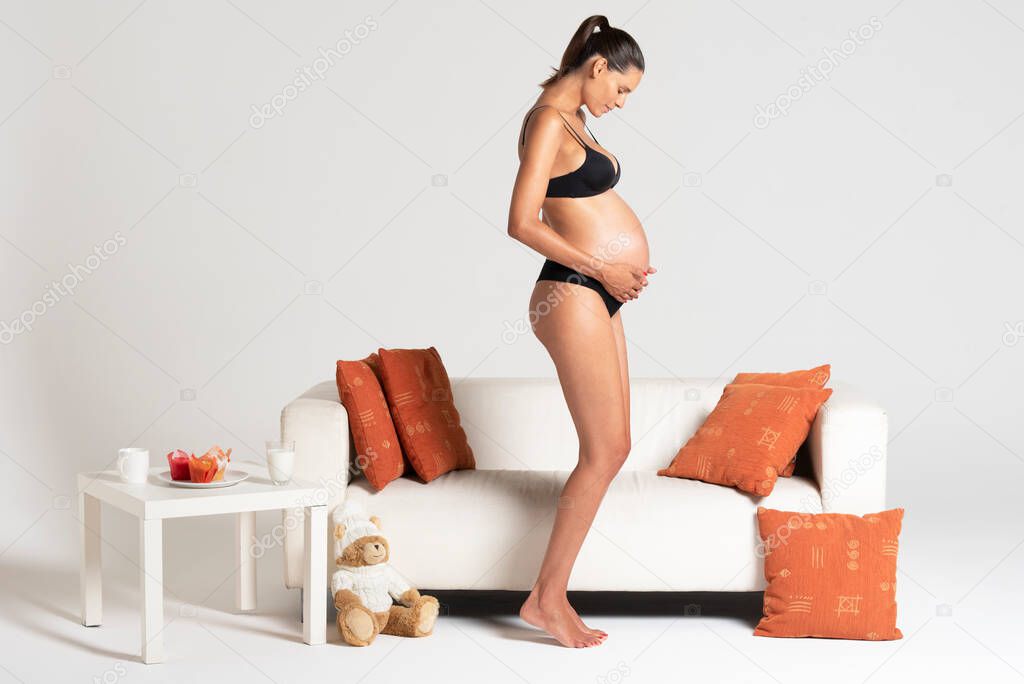 Young beautiful pregnant woman standing close to a white sofa holding hands on her belly dressing underwear