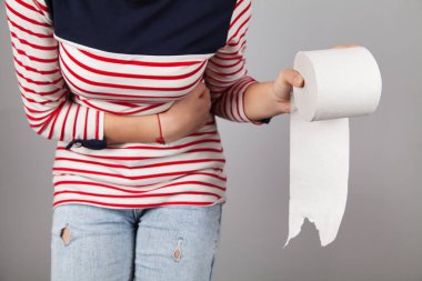 Karın ağrısı çeken genç bir kadın. Kadının elinde tuvalet kağıdı var. Gri arkaplan.