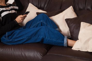 Oturma odasındaki kanepede rahat bir evde akıllı telefon kullanan genç bir kadın. Kanepede deri yastıklar var..