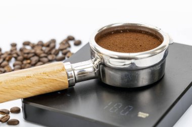İnce öğütülmüş kahveyle dolu bir iskele filtresi, beyaz bir yüzeye yayılmış kavrulmuş kahve çekirdekleri ile 118.0 gram gösteren dijital bir kahve ölçeğinde oturur..