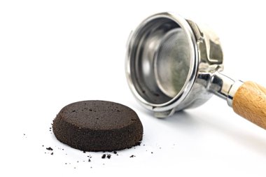 Kullanılmış espresso kahve telvesi beyaz arka planda ahşap saplı paslanmaz çelik bir portafilterin yanında kahve demleme sürecini resmediyor..