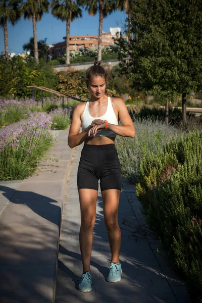 Sporty Caucasian woman looking at her smartwatch after running. Full body frontal photo
