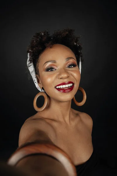 Girl with black skin and curly hair taking a selfie while smiling, made up and dressed in a black shirt, wooden accessories.