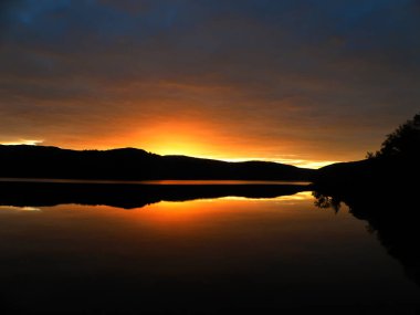 Temmuz sonlarında Murmansk şehrinde yaz günbatımı. Suyun üzerindeki karanlık dağlar, sarı günbatımı, sudaki yansıma, sessiz ağaçlar. Murmansk bölgesi, Rusya.