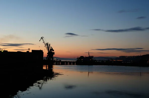 Murmansk à noite, as luzes da cidade são refletidas na baía e os navios atracados no porto. Foto noturna, o contorno do guindaste e do navio no escuro com reflexo na água. — Fotografia de Stock