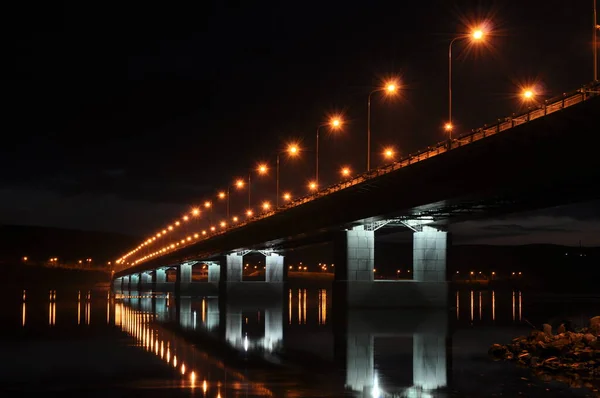 Vista noturna da ponte sobre o rio Kola. A cidade de Murmansk. Uma longa ponte com lanternas iluminadas refletindo na água. Murmansk, Rússia — Fotografia de Stock